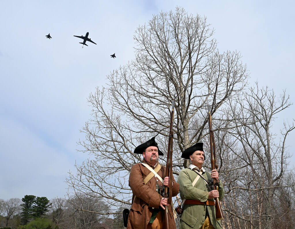 Two F-15’s and a tanker fly over Minute Man as re-enactors Gary Babin and Dan Robson of the 6th Middlesex Regiment stand at attention at the conclusion of the 250th anniversary Patriots Day North Bridge ceremony. Photo: Ken McGagh/The Acton Bridge
