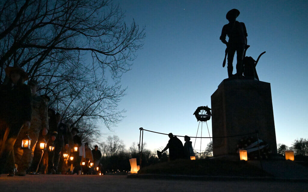 A lantern-light procession goes by the Minute Man statue before a recitation of the names of soldiers from both sides who gave their lives on April 19, 1775, during the eighth annual Patriot Vigil at Minute Man National Historical Park in Acton, April 17, 2025. The path from the North Bridge Visitor Center to the North Bridge was lighted with luminaries to honor those who lost their lives that day. Ken McGagh for The Acton Bridge