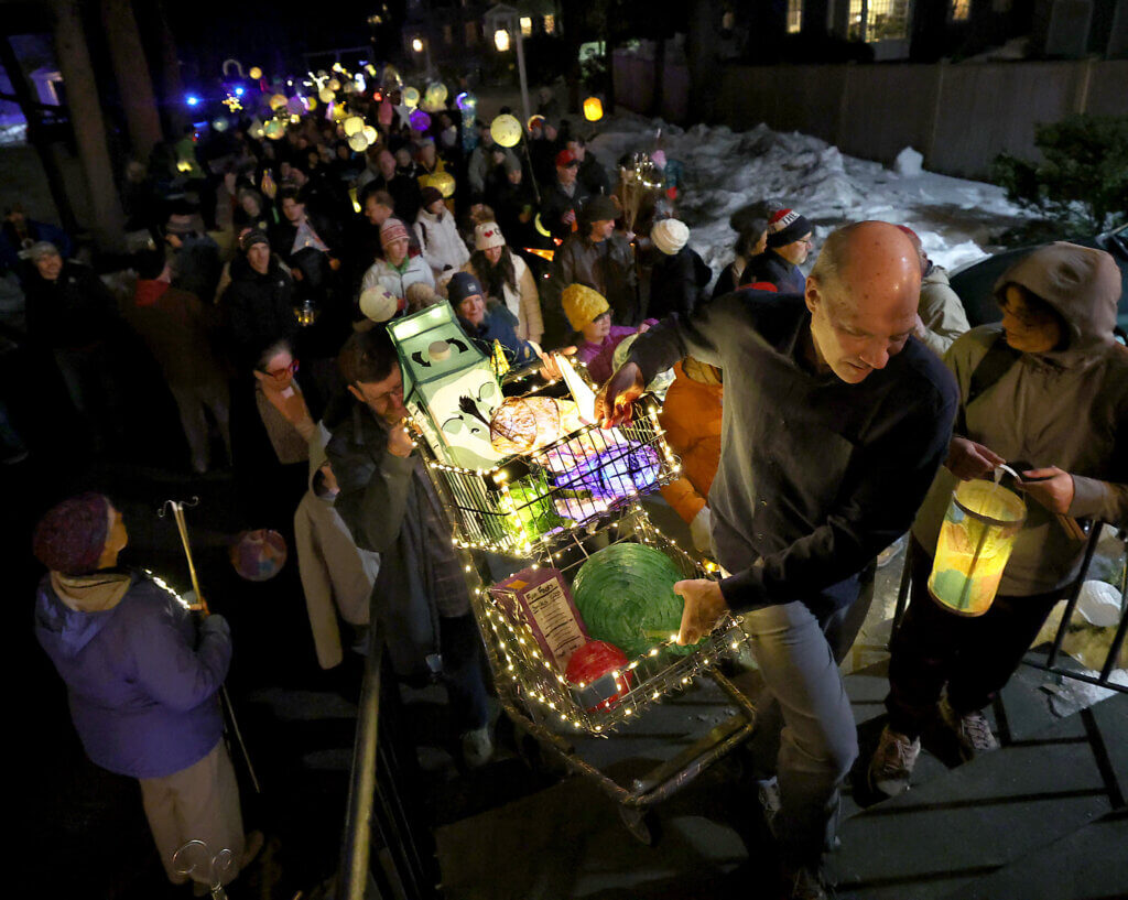 A shopping cart lantern, a collaborative effort by several Arts for All artists and one of the highlights of the inaugural Acton Lantern Parade, is carried into the Scout House.  Adam Stark of Debra’s Natural Gourmet led a team of children pushing the cart through Acton. 