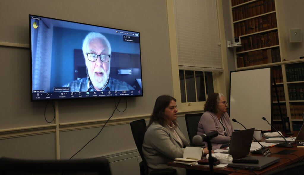 Select Board member Paul Boehm speaks via video in favor of the Holocaust Remembrance Alliance (IHRA) definition of antisemitism as Select Board Chair Mark Howell looks on during the 2026 Town Caucus at the Town House on Tuesday, January 27, 2026. Ken McGagh/The Acton Bridge