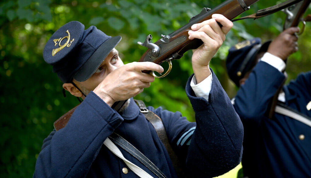 Emmett Bell-Sykes of Boston, portraying Sgt. William H. Carney, the first Black soldier to receive the Medal of Honor, demonstrates how he fires his rifle during a living history Civil War era encampment with re-enactors of the 54th Massachusetts Volunteer Infantry, the first officially recognized Black regiment in the Union Army, at the Robbins House in Acton on Saturday, July 26, 2025. Ken McGagh/The Acton Bridge