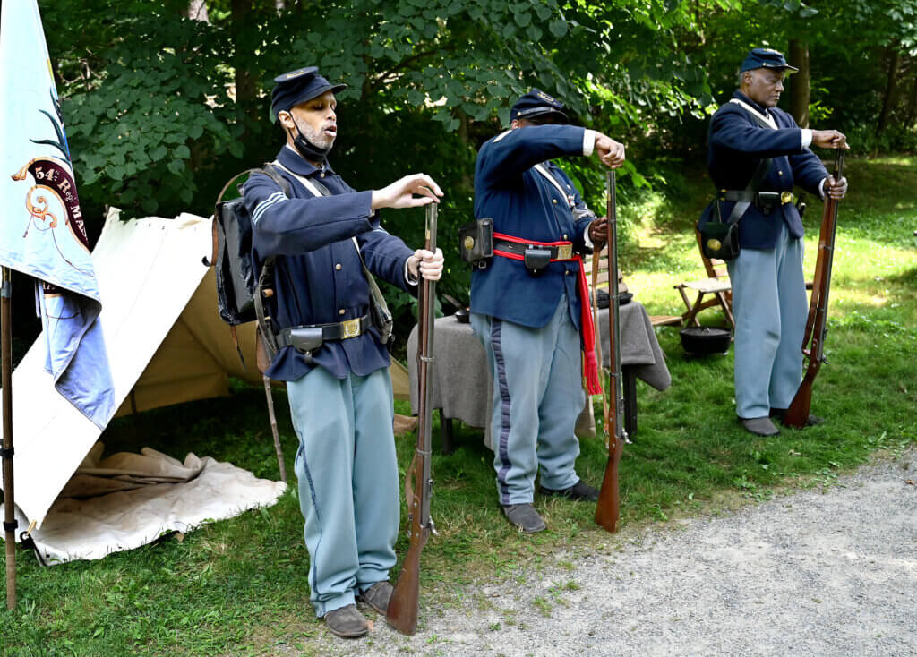 Emmett Bell-Sykes of Boston, left, Gerard Grimes of Boston and Joe Zellner of Bedford, re-enactors of the 54th Massachusetts Volunteer Infantry, Company A, demonstrate how they load their rifles during a living history Civil War era encampment at the Robbins House in Acton on Saturday, July 26, 2025. Ken McGagh/The Acton Bridge