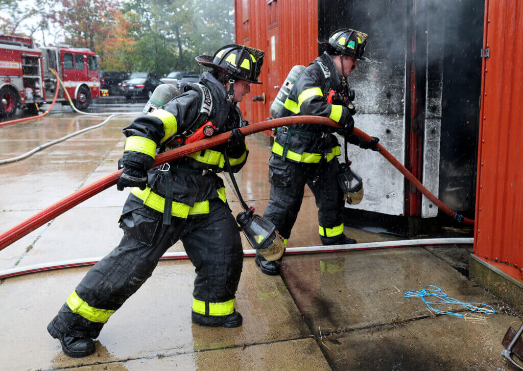Acton firefighters Elsa Heil, left, and Keegan Byrnes load hose into a burning building during “refresher” training session at the Worcester Fire Department Training Center on Monday, September 27, 2026. Ken McGagh/The Acton Bridge