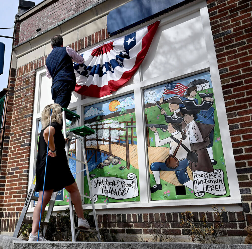 John Cadigan and Raeann DeRienzo of Barrett Sotheby’s International Realty hang patriotic  bunting above a brand new Concord250  window mural  at 33 Walden Street in Acton Center on Friday, April 4, 2025.  The painting was created by a three-person crew led by muralist Maura Gorman on Wednesday.  Passersby are encouraged to pose for a selfie and use the hashtag  #bsir250 for the chance to win $250.00. The artist has hidden some “250’s” throughout the art work as well.   Ken McGagh/The Acton Bridge