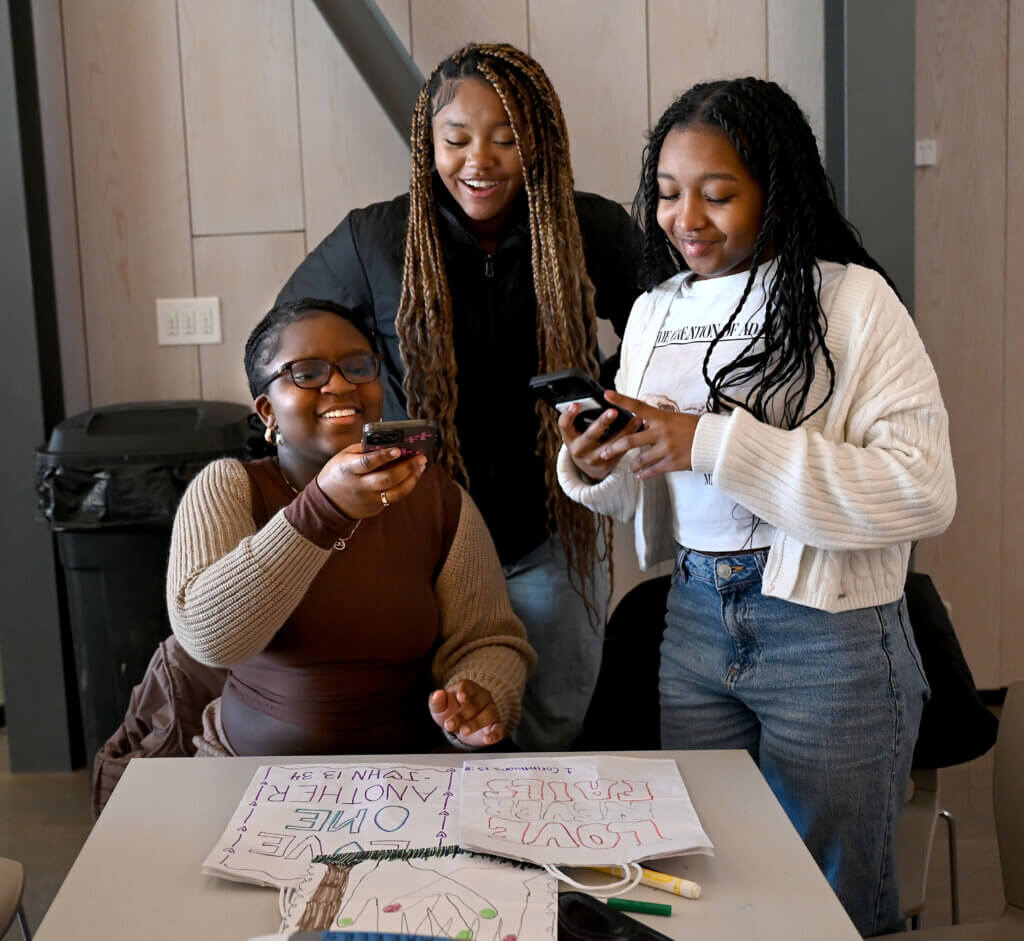 Acton Academy freshmen Melissa Asare, Ray’Hannah Mendez and Avery Barkley take a cell phone picture after decorating bags that will hold meals and snacks for local children at the Acton Museum on Monday, January 20, 2025. The Martin Luther King Day of Service activity was hosted by the Acton Museum Contemporary Leaders in partnership with Open Table. Ken McGagh/The Acton Bridge
