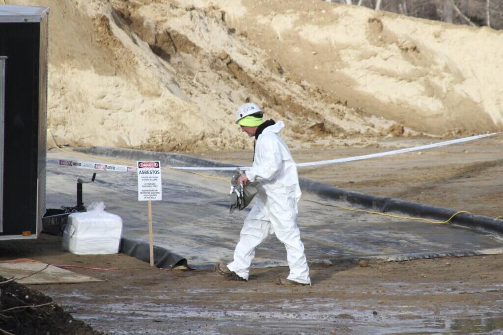 A worker in protective gear walks past a sign warning about asbestos in soil at the site of a planned West Acton subdivision. Photo: Dakota Antelman/The Acton Bridge