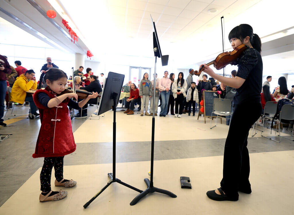 Summer Chen, 5, of Acton, left, her sister Hannah Chen, 13, play violin n the cafeteria during the third annual Lunar New Year Celebration at Acton-Carlisle High School on Saturday, February 7, 2026. Two sisters also performed later on the main stage. Ken McGagh/The Acton Bridge