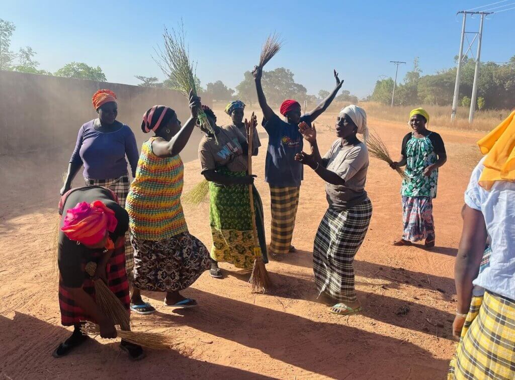 Women dancing with brooms during a community cleanup exercise. Photo by Mikaela Smith