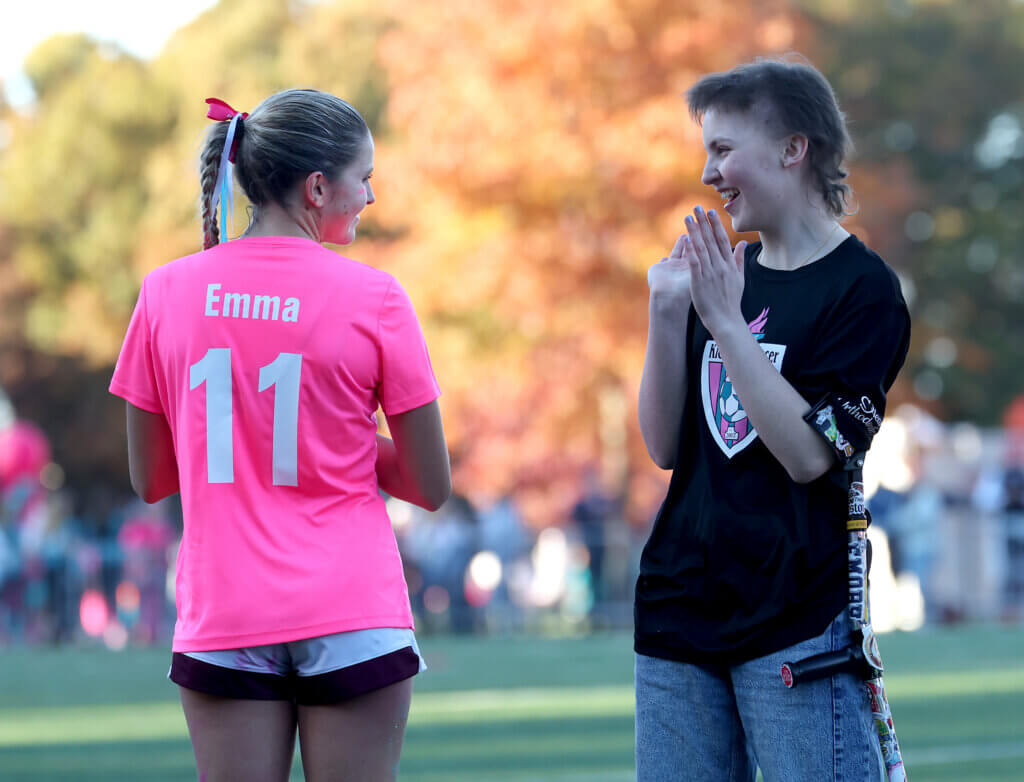 During a halftime ceremony, Acton-Carlisle co-captain Emmy O’Donnell, left, shows the crowd that her jersey honors classmate Emma Robertson, right, a brain cancer survivor.