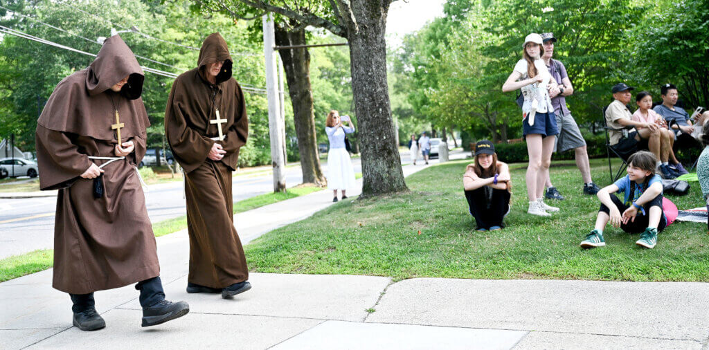 A pair of friars catch the eye of the audience as they walk on the Acton Free Public Library sidewalk during The World’s A Stage Players production of William Shakespeare’s “Henry IV, Part I” on Saturday, July 26, 2025. Two more outdoor performances are scheduled for August 2 and 3 at the library. Ken McGagh/The Acton Bridge