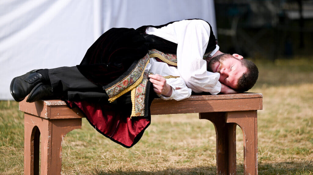 Alex Wersted, as Prince Hal, sleeps on a bench in the opening scene of The World’s A Stage Players production of William Shakespeare’s “Henry IV, Part I” on the Acton Free Public Library lawn on Saturday, July 26, 2025. Ken McGagh/The Acton Bridge