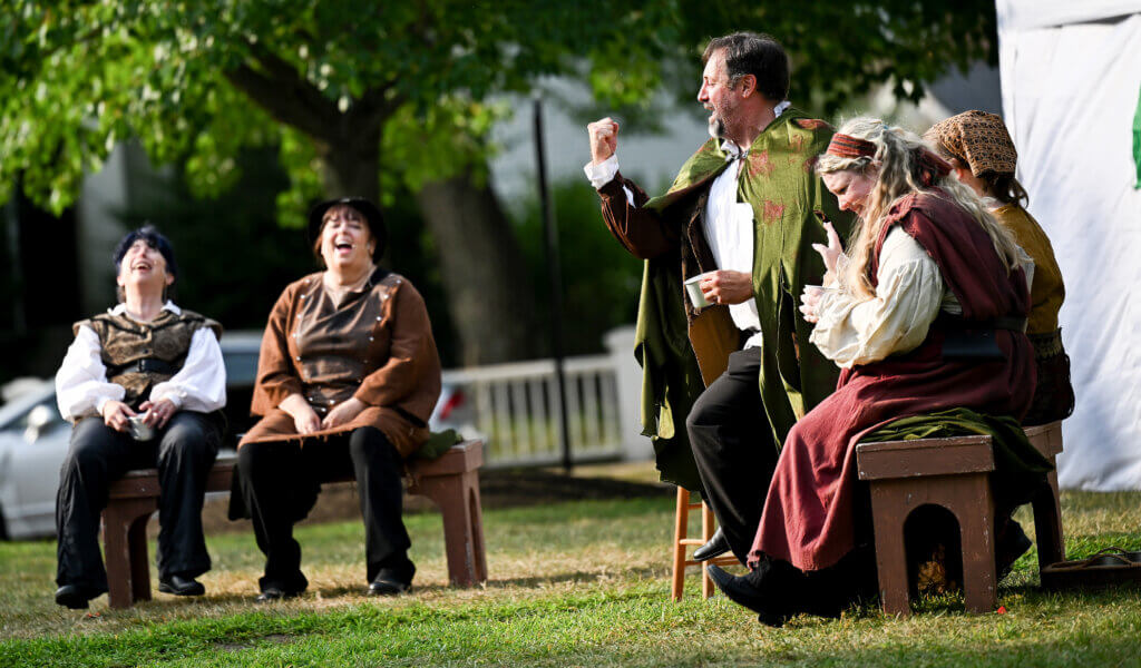 Mike Haddad, as Prince John of Lancaster and other Acton Players actors laugh during The World’s A Stage Players production of William Shakespeare’s “Henry IV, Part I” on the Acton Free Public Library lawn on Saturday, July 26, 2025. Haddad is also the production’s director. Ken McGagh/The Acton Bridge