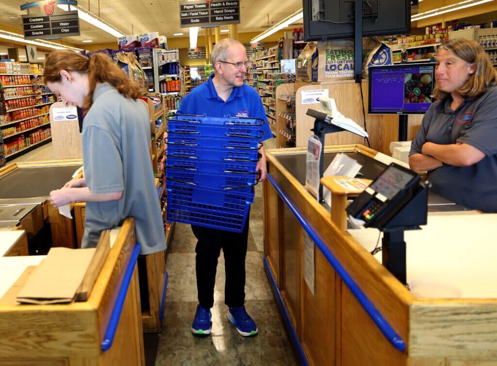 Inferrera carries baskets through a checkout aisle between cashiers Daniel Edelman, left, and Erica Cyr. Photo: Ken McGagh/The Acton Bridge