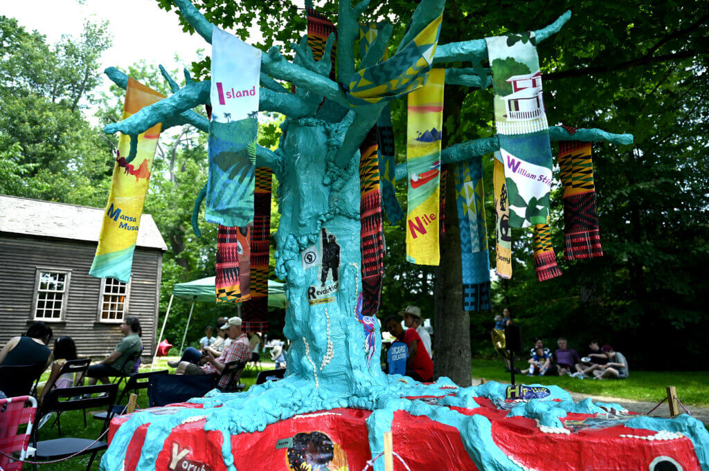 A tree sculpture by artists Char Jere and Meaghan Elyse, made of plaster and fabric and based on the picture book “Ayo’s Adventure: Across the African Diaspora from Afro to Zulu,” sits behind people enjoying music at The Robbins House.
