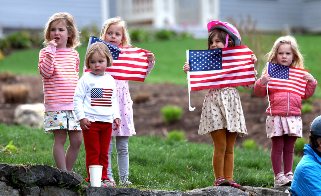 Acton residents Josie Lockwood, 5, left, Marian Ott, 2, Margot Clapp, 4, Weezy Terell, 5, and Abigail Terell, 3, hold American flags as the Acton Patriots Day parade passes their homes on Monday.  
