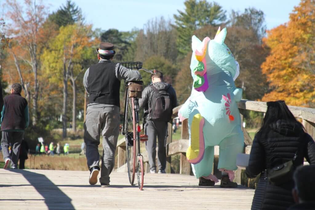 Chris Beatrice of Maynard pushes his penny-farthing bicycle past a person in a unicorn suit. Photo: Dakota Antelman/The Acton Bridge