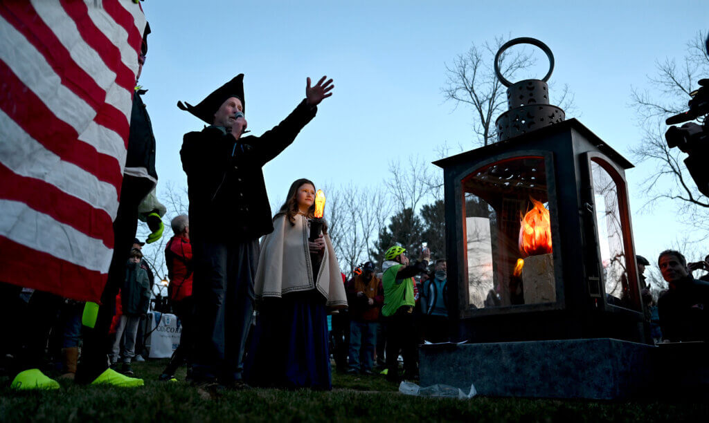 Richard Fahlander of Art for All, left, speaks to a crowd gathered on the Acton Museum lawn as Liviana Stevens, 11, holds an LED torch after the ceremonial lighting of the Concord250 Lantern. Liviana, of Littleton, also sang the national anthem. She is the granddaughter of Priscilla and Jonathan Stevens of Acton. Photo: Ken McGagh/The Acton Bridge
