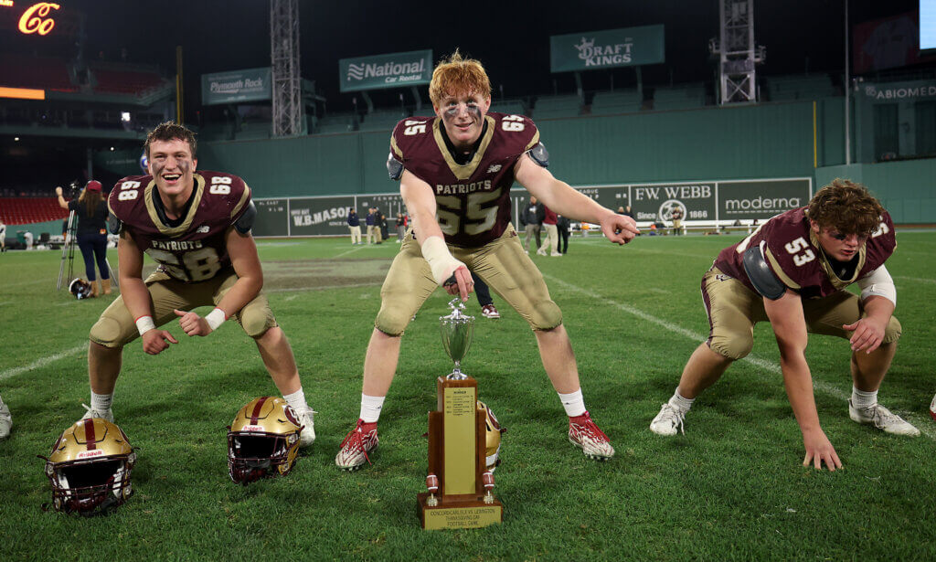 Acton-Carlisle football players Owen Root, left, Tyler Colgan, and Eric Nick form a line of scrimmage with the Patriots vs. Minutemen Thanksgiving Football trophy after CC’s 32-0 win at Fenway Park on Wednesday, November 25, 2025. Photo: Ken McGagh/The Acton Bridge