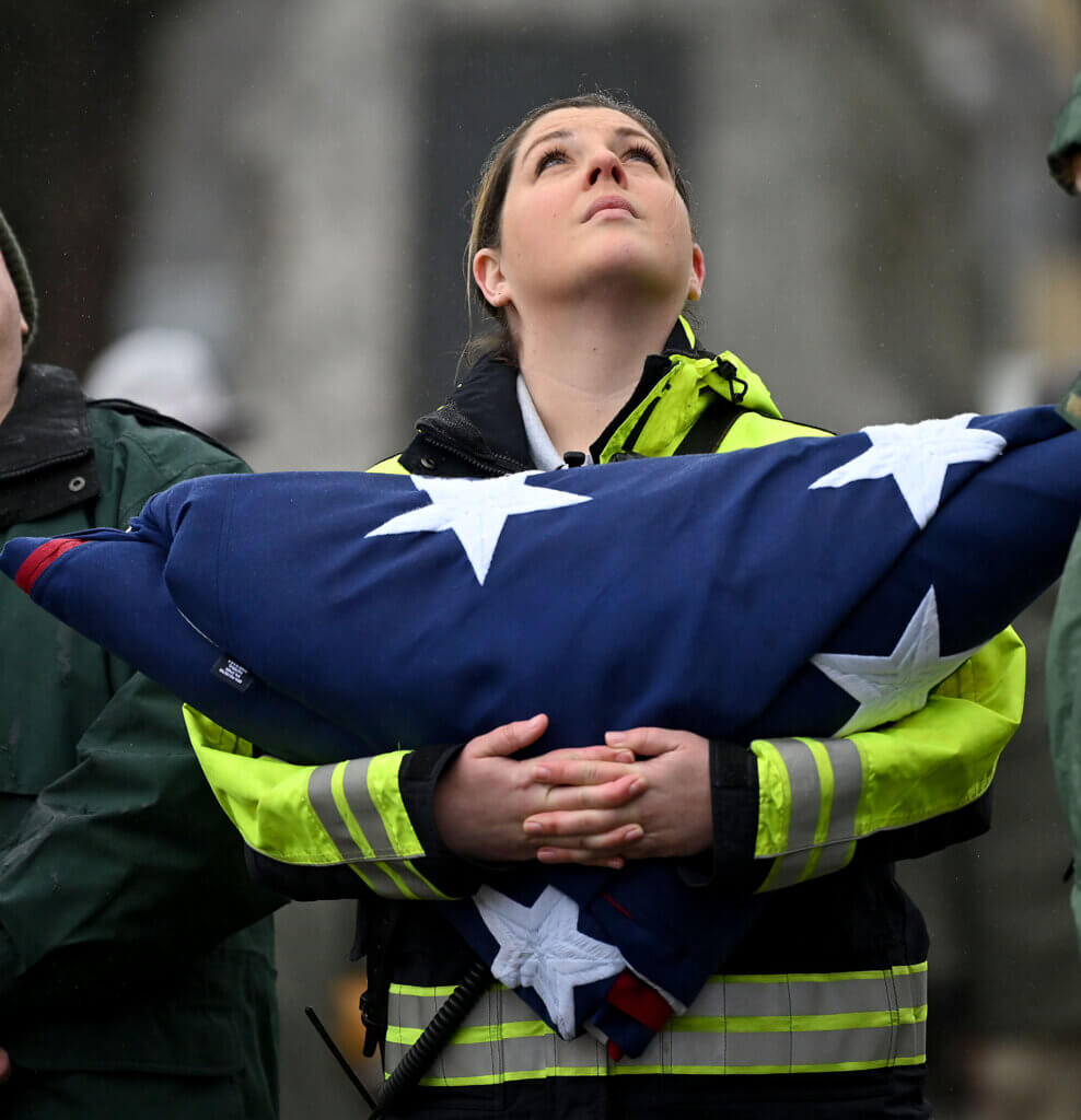 Acton Fire Department Lt. Lindsay McGloughlin, holding the town’s new 15’x25’ American flag, looks up as fellow firefighters take down the old Old Glory during a flag-raising ceremony in Acton Center on Saturday, April 12, 2025. Ken McGagh/The Acton Bridge