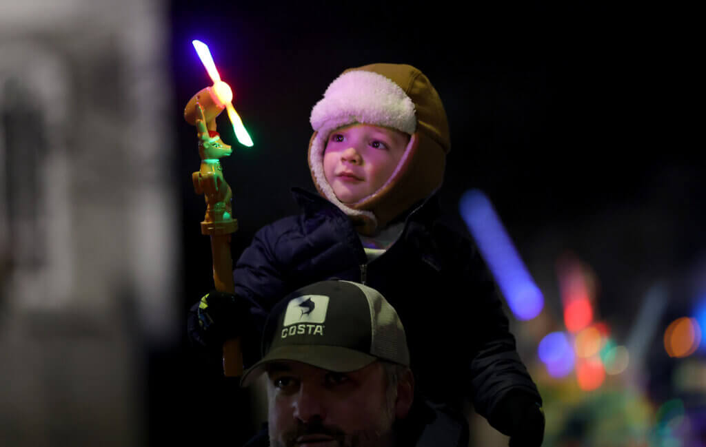 Jack Igoe, 3, of Lexington, plays with his holiday-theme light-up spinning wand while sitting on the shoulders of his fathter, John Igoe, after Santa Claus turned the switch on the town’s Christmas tree in Monument Square on Sunday, December 7, 2025. Igoe said that his parents and cousins live in Acton. Ken McGagh/The Acton Bridge
