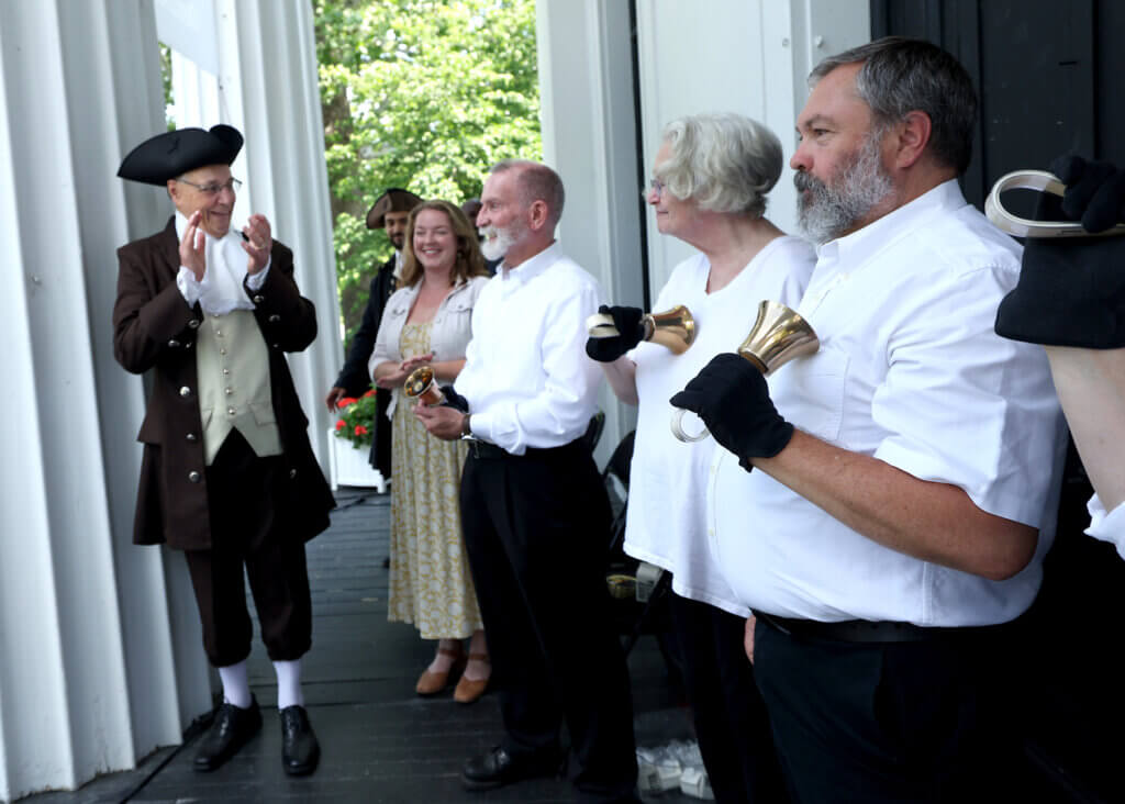 Moderator Tom Wilson of the Wright Tavern Legacy Trust, left, applauds bell ringers from the Trinity Episcopal Church at the conclusion of “Declaring Independence Then and Now In Acton” on the steps of the First Parish in Acton on Saturday, July 11, 2026. Ken McGagh/The Acton Bridge