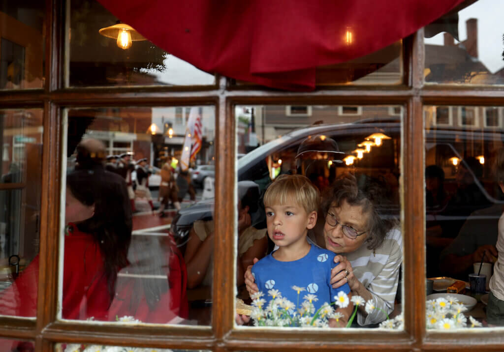 Vicky Slavin of Lincoln and her grandson, Leo DeRanieri, 4, watch from Main Streets Market & Cafe as Monday’s parade goes by.  Ken McGagh/The Acton Bridge 
