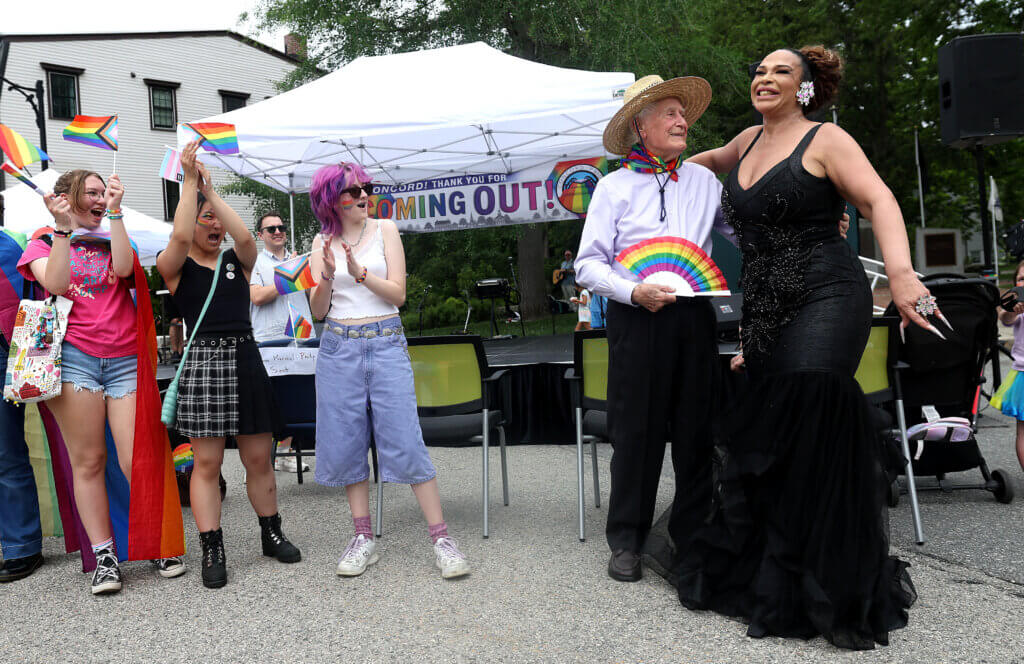 Drag queen Jazmine Skies, right, hugs Philip B. Dee Jr., parade Grand Marshal, as a group of teenager cheer on, during Acton Pride Festival Mizery Loves Company live show in Monument Square on Saturday, June 6, 2026. Ken McGagh/The Acton Bridge