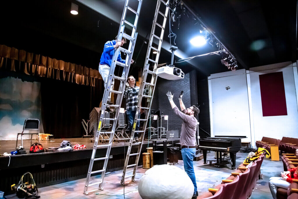 People affiliated with the Acton Scout House lower a projector from the ceiling of the Sanborn school auditorium. Photo: Stephen Beyer/The Acton Bridge