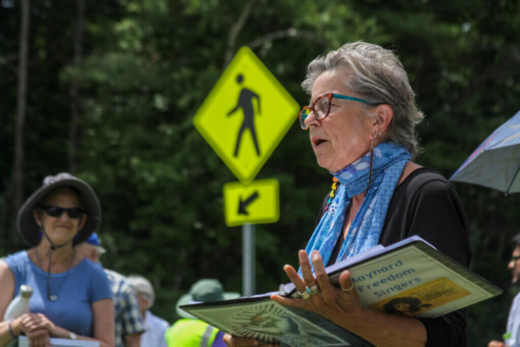 Maynard Freedom Singers leader Sally Sweitzer performs during a protest near the Hanscom Field entrance. Photo: Dakota Antelman/The Acton Bridge