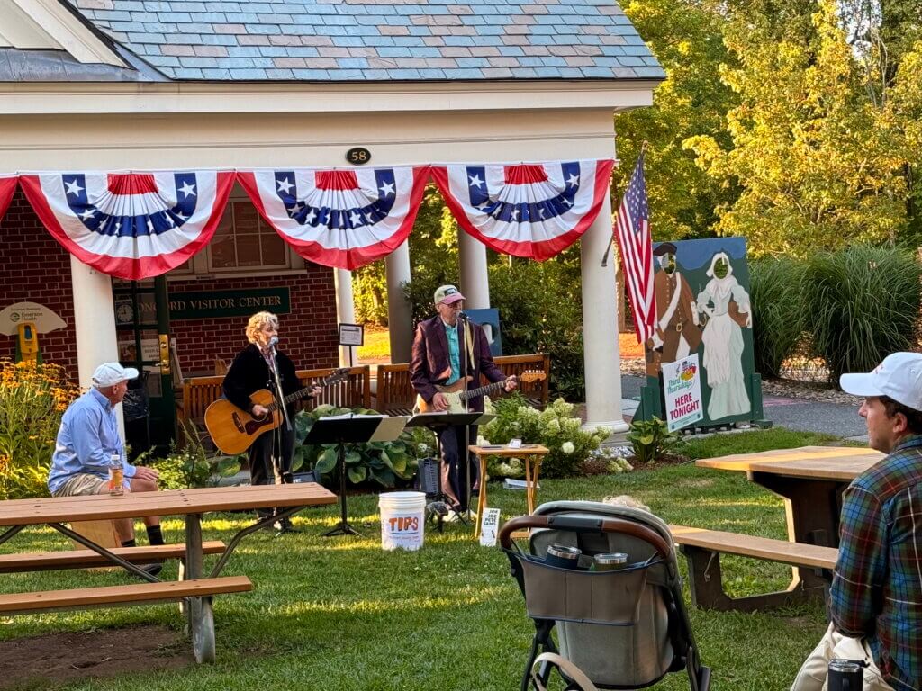 Ruth Rappaport and Joe Pete Wetherbee play roots rock jams in front of the Acton Visitor Center as spectator Rama Valianti joins in on a wooden block. Photo by Elisabeth Weber