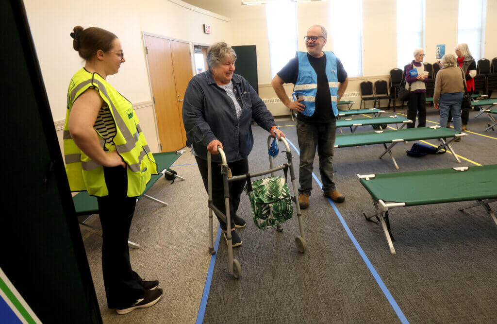 Ione Nauseda, center, acting as a shelter-seeker, is ushered through cots by Erin McMorrow, left, of Acton human services, acting as assistant shelter manager, and Mike Funaiole, right, Acton Public Health Inspector. Photo by Ken McGagh/The Acton Bridge
